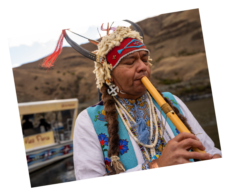 Landing Page A polaroid of JR Spencer, a Nimiipuu storyteller, playing an instrument near the Snake River backed by a rugged landscape.