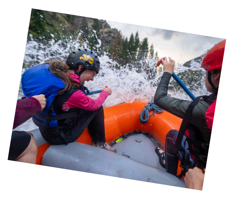 Landing Page A polaroid of a girl in a helmet laughing as her whitewater rafting group hits a big, splashy rapid in their orange raft on the Main Salmon River, with trees ahead.