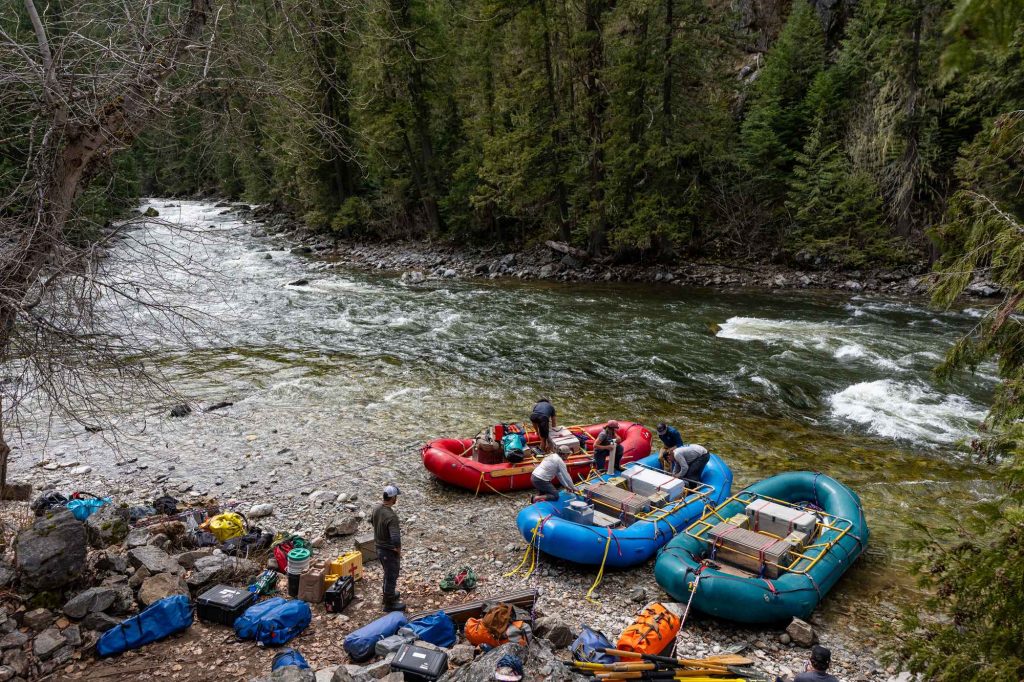 Landing Page Adventurers stand by their rafts, gear spread across riverside rocks, preparing to ride the Selway River.