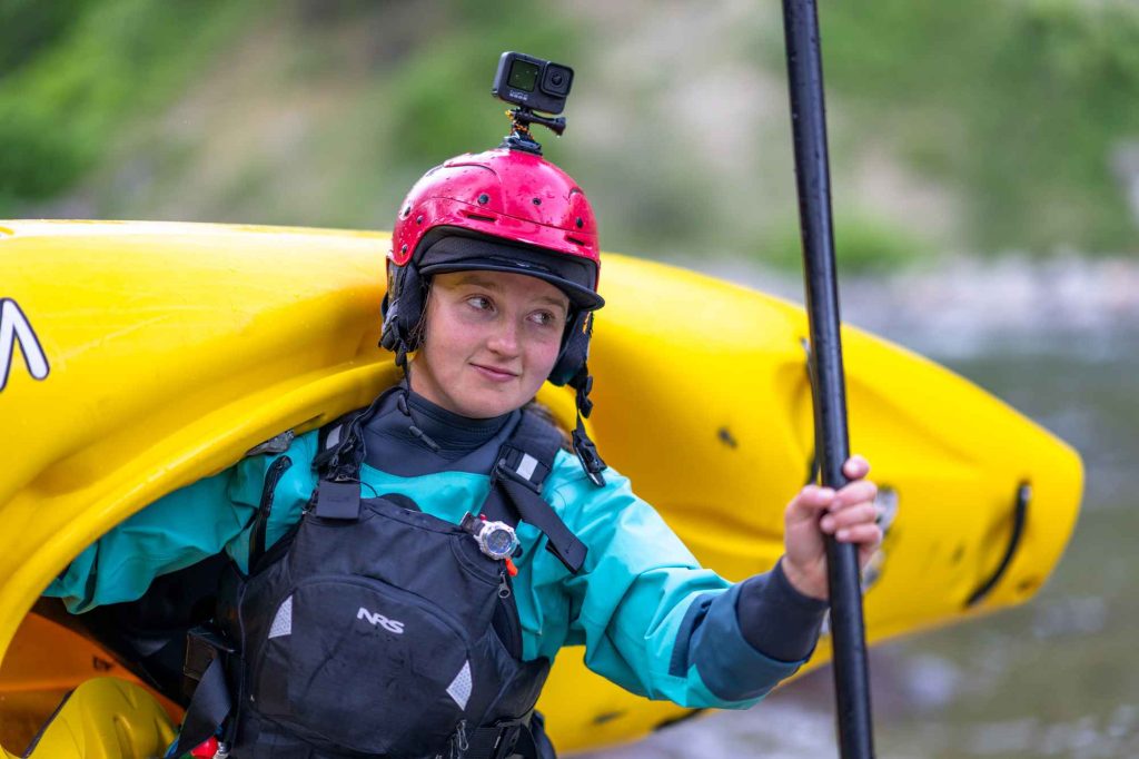 Landing Page A professional kayaker on the Payette River lifts a kayak and raises an oar, wearing a helmet-mounted camera.